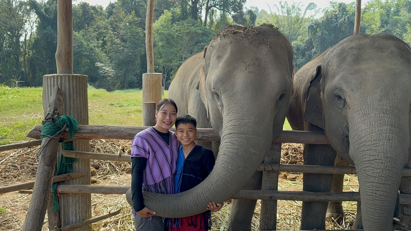 Two smiling young visitors as an elephant curls its trunk around them beside another elephant
