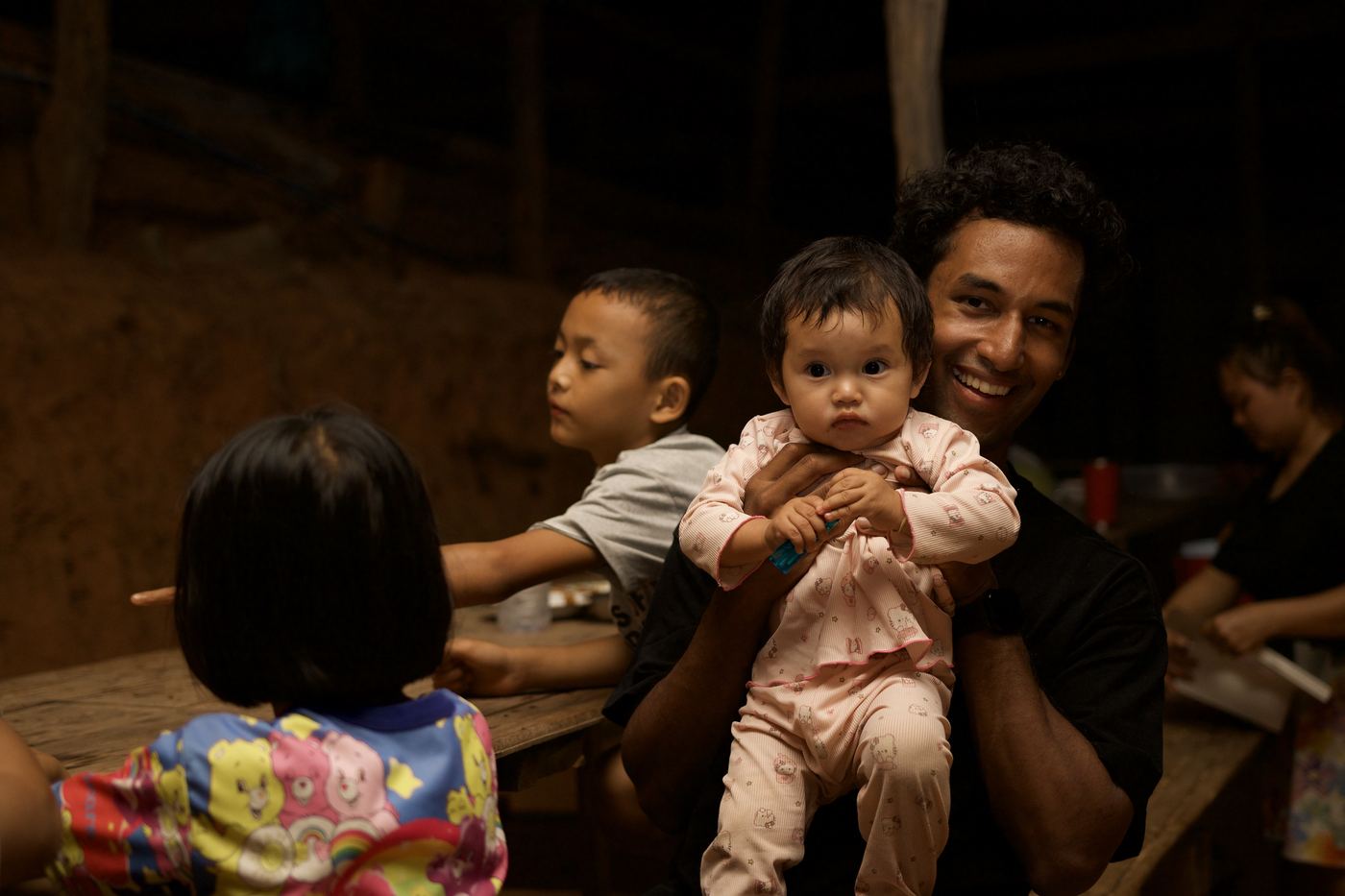 A smiling guest holding a baby while village children gather around a wooden table